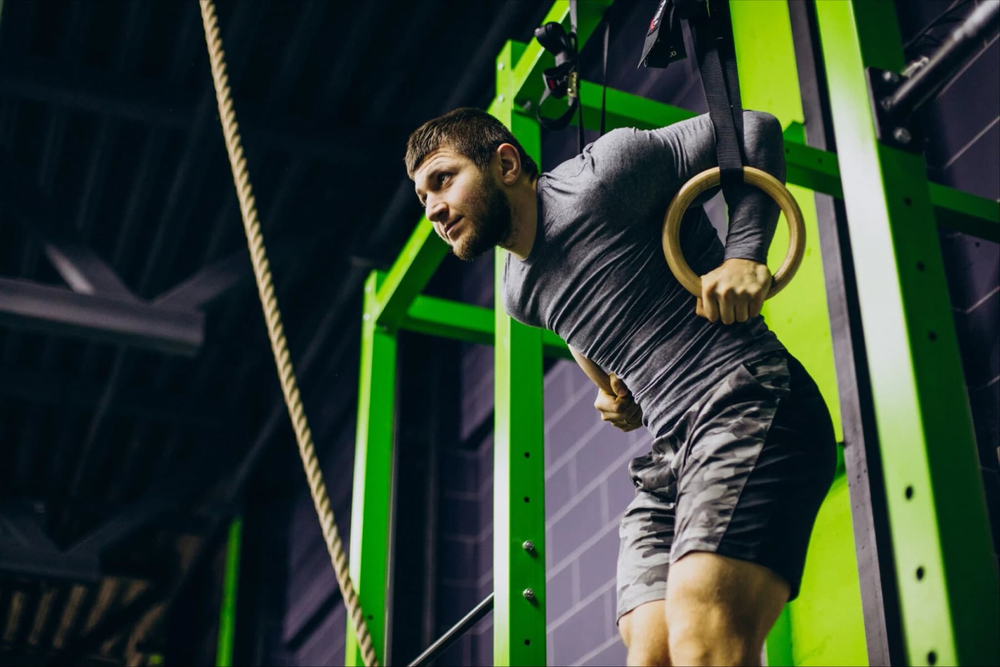 Hombre sosteniendo dos anillos de gimnasia Hombre colgado sosteniendo dos anillos de gimnasia. Su espalda se encuentra levemente inclinada hacia adelante y se observa una cuerda frente a él.