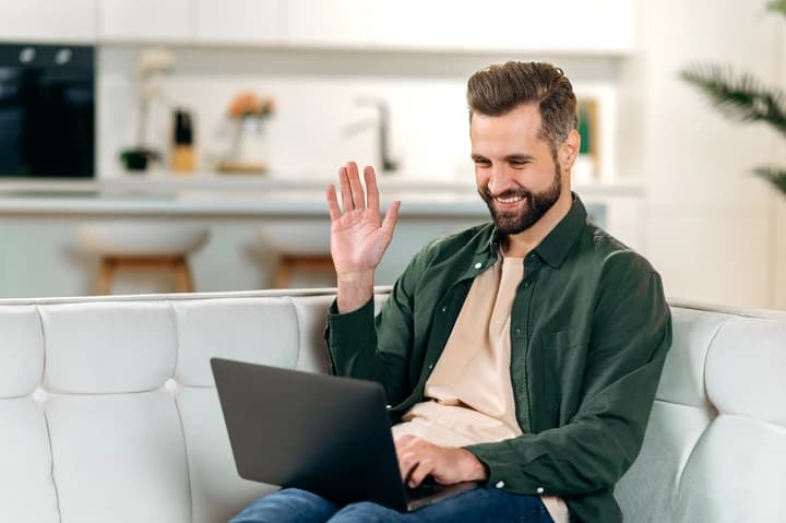 A imagem tem um homem de meia-idade sentado no sofá, sorrindo e acenando para a câmera de um notebook.