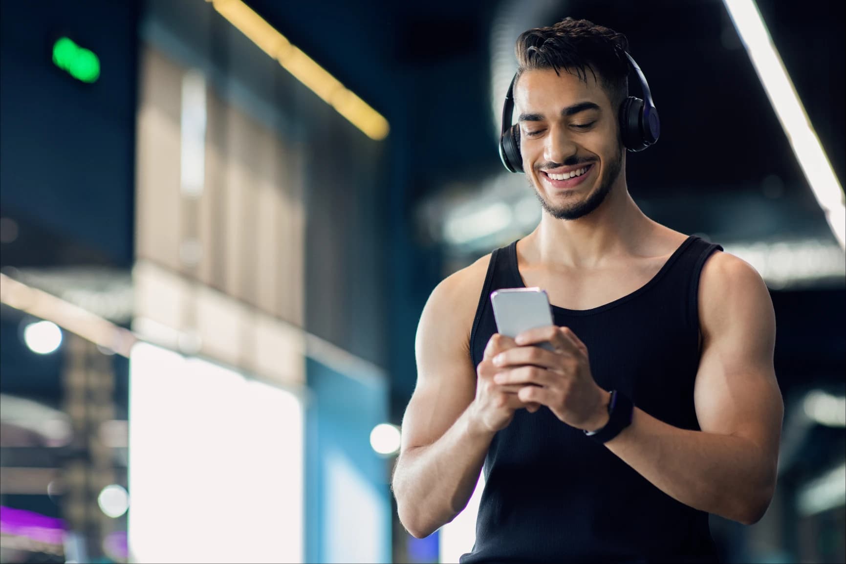 Hombre sonriente con audífonos sosteniendo su celular Hombre con audífonos y reloj, sosteniendo su celular con las dos manos mientras lo observa y sonrie. Al fondo se ven lámparas encendidas y cristales.