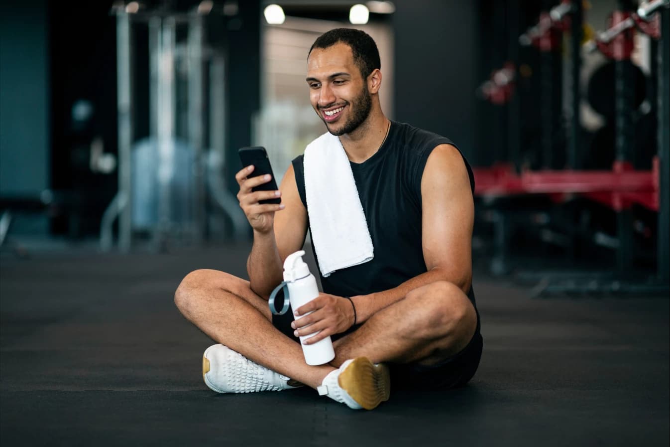 Hombre sentado en un gym con una toalla, agua y su celular Hombre sentado en un gym con los pies cruzados, una toalla, botella de agua y su celular en mano. Al fondo se ven aparatos de ejercicio desenfocados