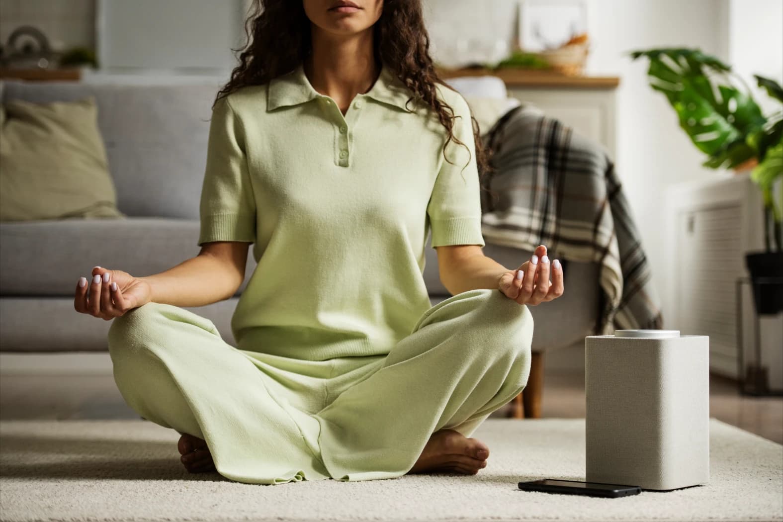 Mujer sentada con las piernas cruzadas meditando Mujer sentada con las piernas cruzadas y sus manos sobre ellas en posición de meditación. Su celular está en frente y al fondo se observa un sillón.