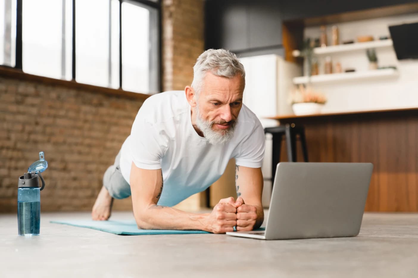 Total Play A imagem tem um homem mais velho treinando. Ele está fazendo o exercício prancha, enquanto olha para o notebook.
