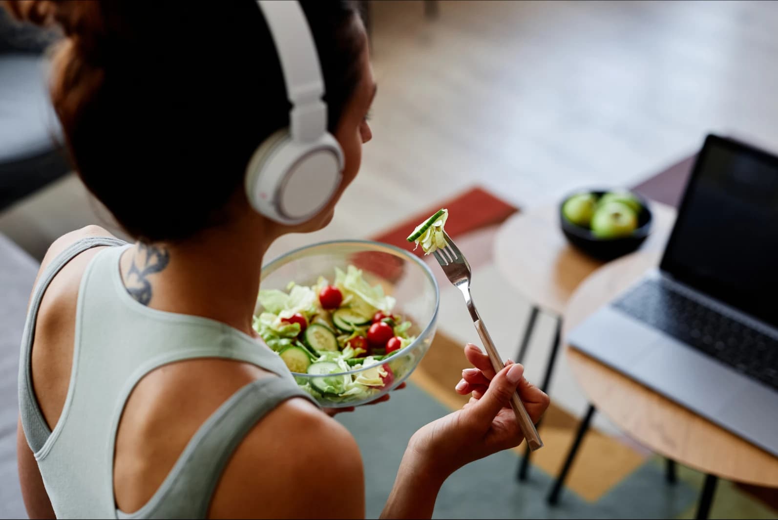 Mujer comiendo una ensalada verde en su casa Mujer con audífonos comiendo una ensalada verde frente a una computadora para mantener una alimentación equilibrada y nutritiva.