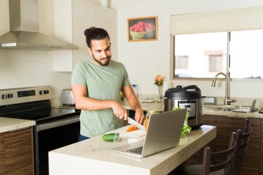 Homem cozinhando pelo Total Nutri. A imagem tem um homem mais jovem cozinhando. Ele está vendo a receita pelo notebook, enquanto corta legumes.