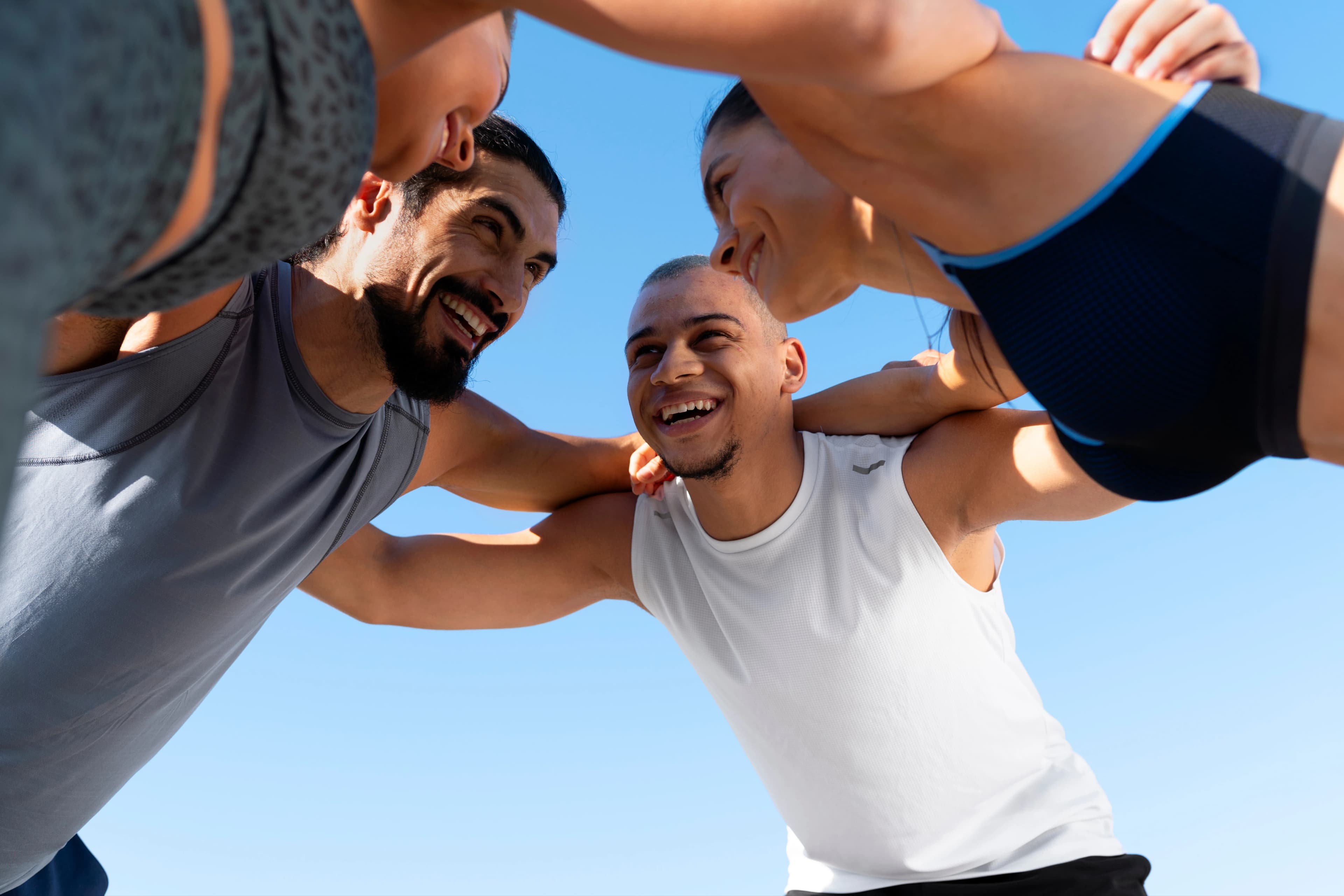 Cuatro personas abrazadas con ropa deportiva Dos mujeres y dos hombres abrazados con ropa deportiva, están ligeramente inclinados y formando un círculo con sus brazos. De fondo se ve el cielo.